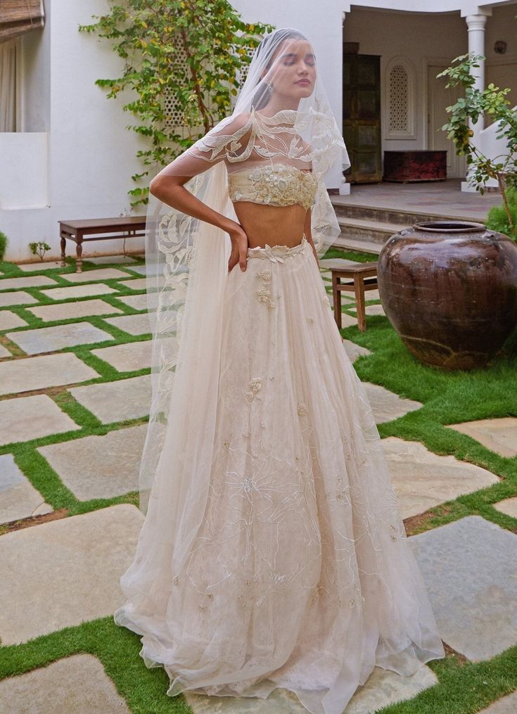 Elevate your style with the stunning Ivory Floral Embroidered Lehenga Set, a breathtaking blend of traditional elegance and modern sophistication. Crafted from luxurious organza, this ivory lehenga is layered with soft silk and French lace, creating a rich, textured base. Intricate tone-on-tone threadwork and zardosi embroidery enhance the fabric, while hundreds of shimmering, hand-cut organza flowers are scattered throughout, adding a whimsical, ethereal charm. The waistline, adorned with delicate floss embroidery and hand-appliquéd beads, exudes opulence and grace. Paired with a strapless crop blouse, featuring sparkling beads and intricately hand-cut organza flowers. With peek-a-boo lace details and an underlayer of fine French lace and silk, the blouse is both romantic and contemporary White Wedding Lehenga, Ivory Lehenga, Zardosi Embroidery, Organza Skirt, Organza Flowers, Embroidered Lehenga, Indian Wedding Wear, Wedding Lehenga, Lace Embroidery