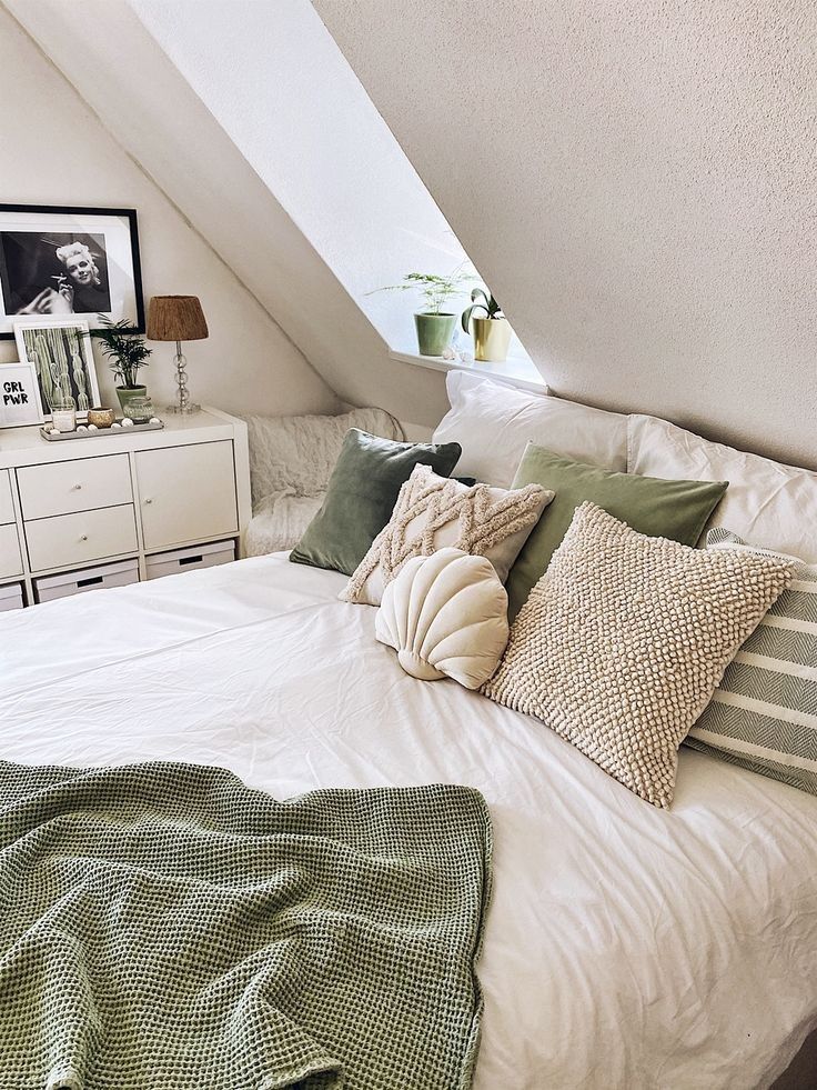 a white bed topped with lots of pillows under a slanted ceiling next to a dresser