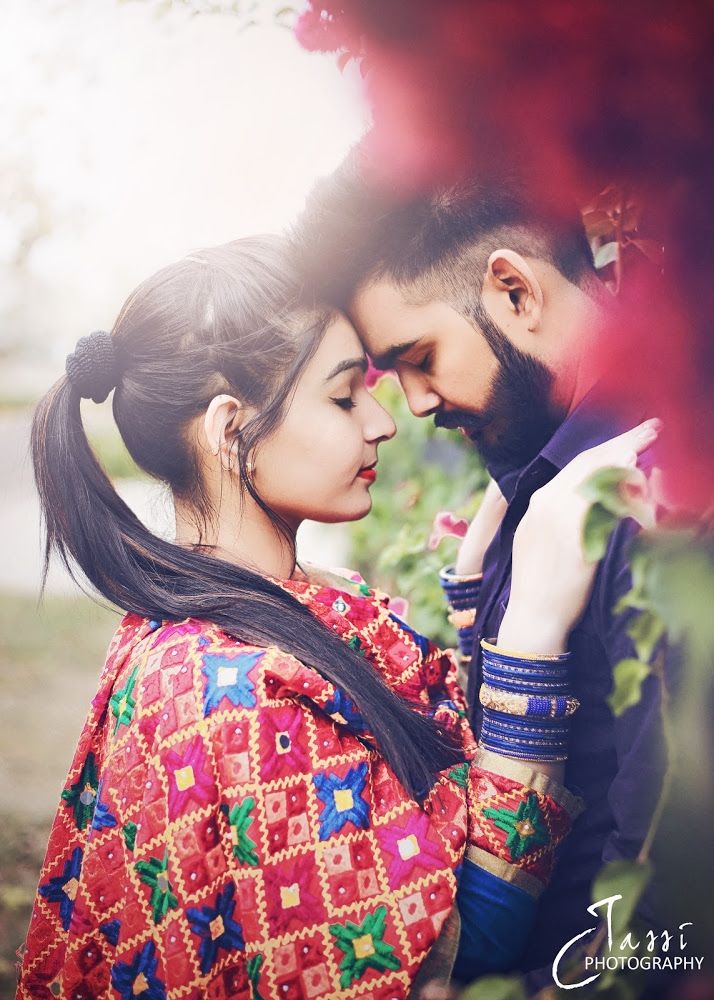 a man and woman standing next to each other in front of some red flowers with their arms around each other