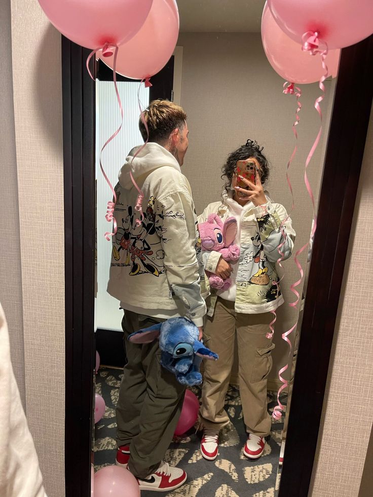 two people standing in front of a mirror with balloons on the floor and one person holding a stuffed animal