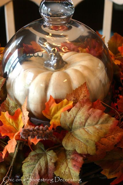 a glass covered dish with pumpkins and autumn leaves on the table in front of it