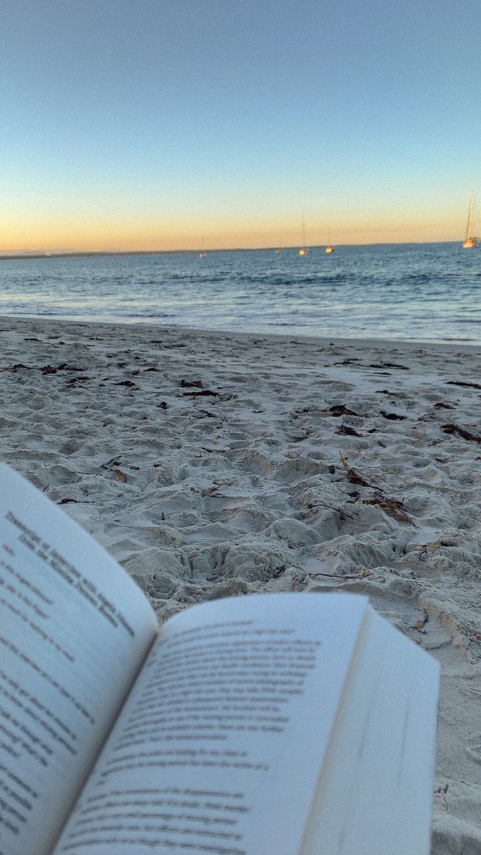 an open book sitting on top of a sandy beach next to the ocean with sailboats in the distance