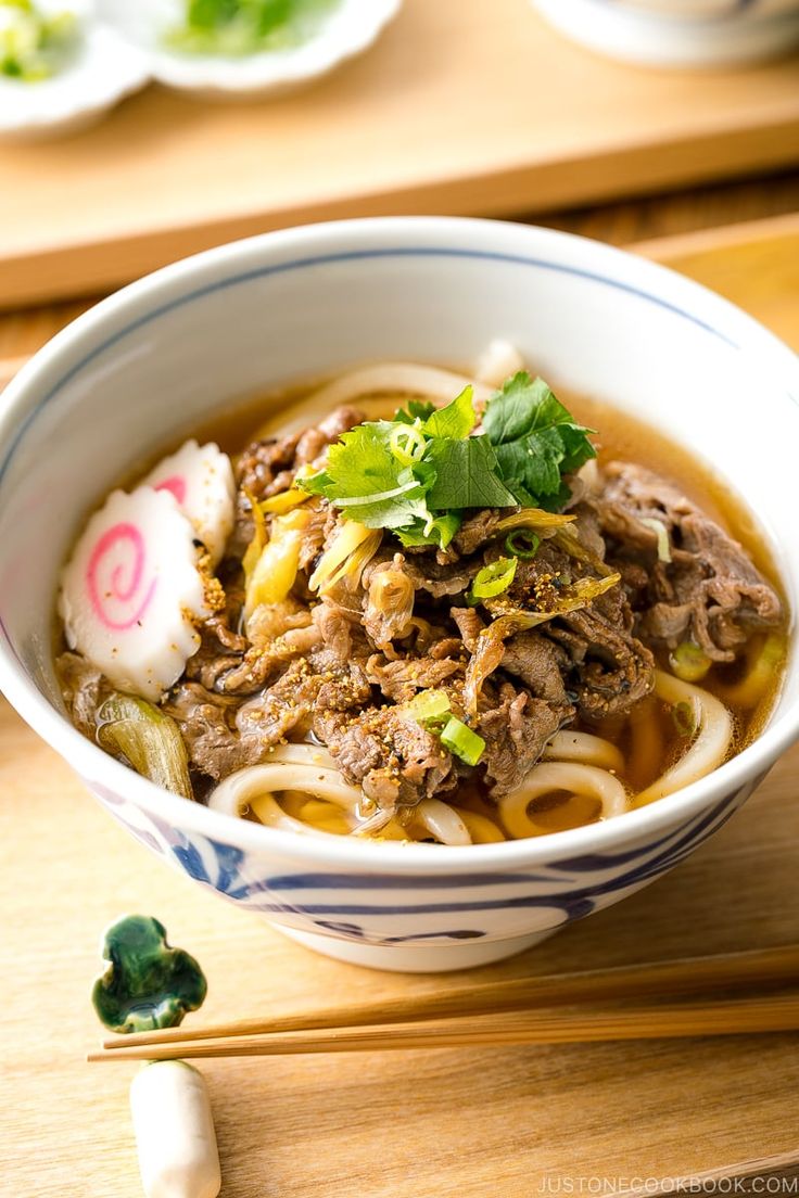 a bowl filled with noodles and meat on top of a wooden table next to chopsticks