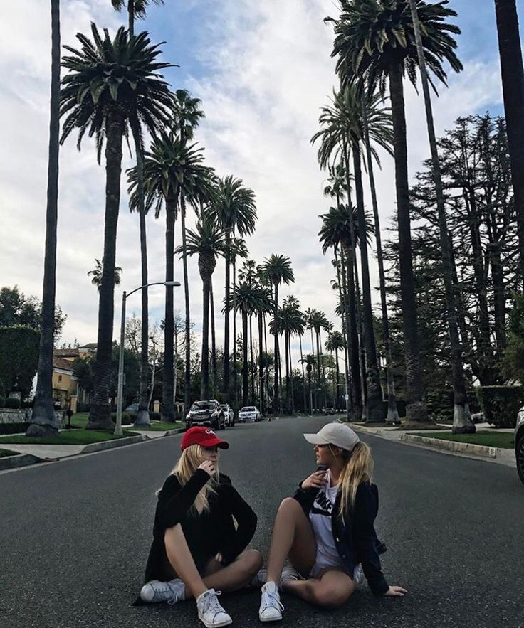 two young women sitting on the side of a road next to tall palm tree lined street