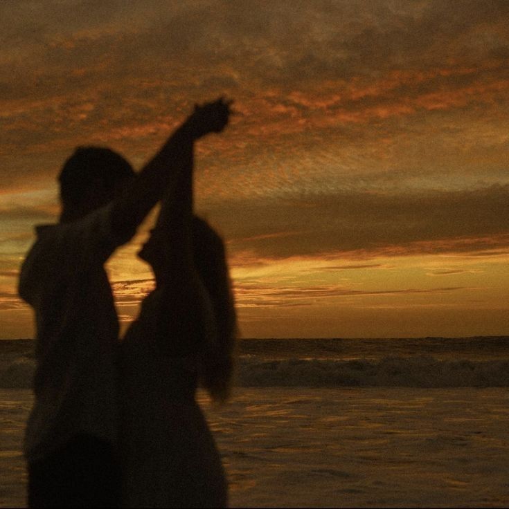 two people standing on the beach at sunset with their arms in the air and one person reaching for something