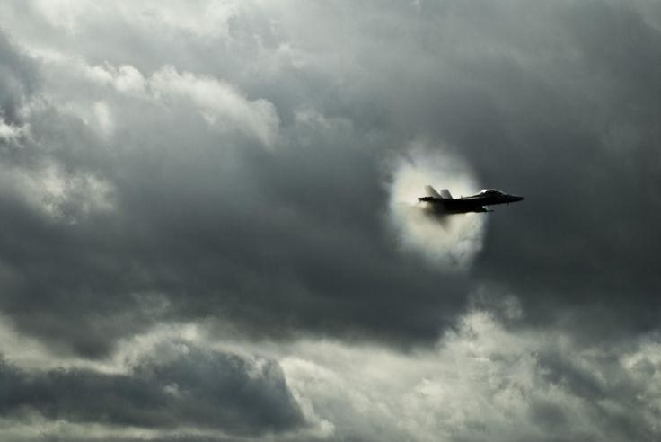 a fighter jet flying through a cloudy sky with the sun peeking out from behind it