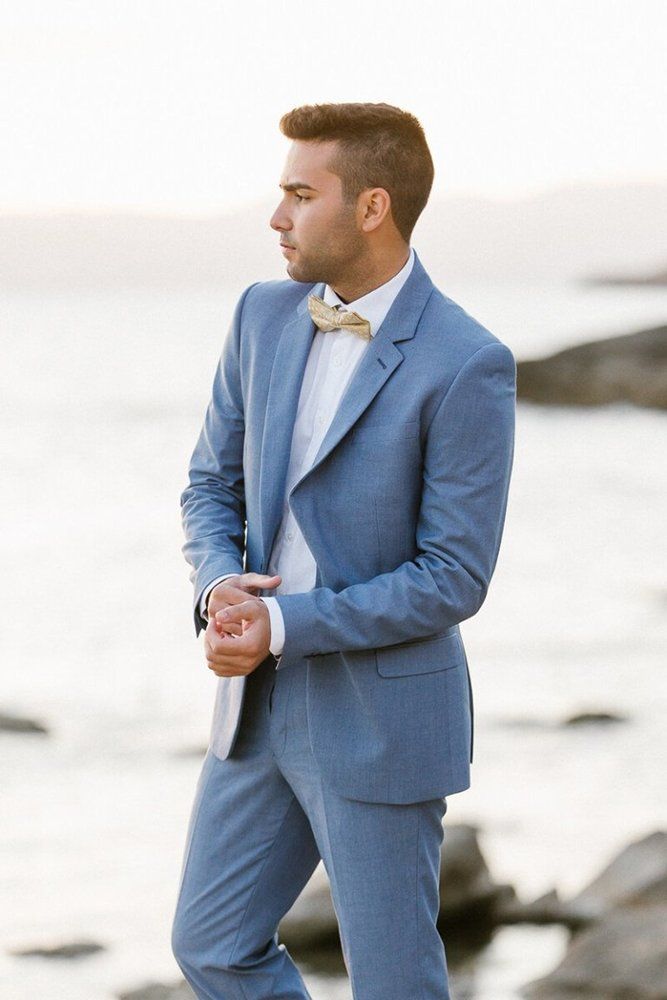 a man in a blue suit and bow tie standing by the ocean with his hands in his pockets