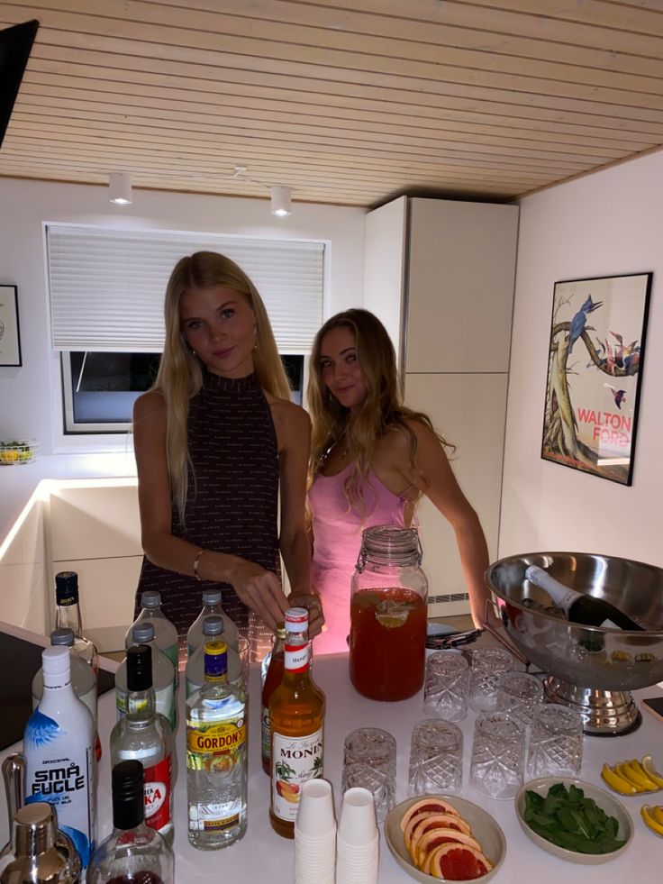 two women standing in front of a table full of drinks and condiments on it