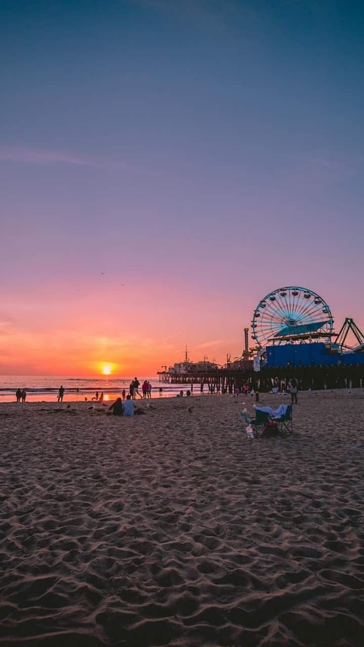 the sun is setting at the beach with people sitting on the sand and an amusement park in the background