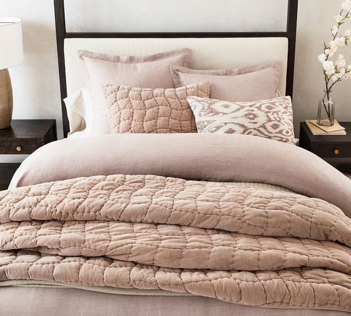 a bed that has some pillows on top of it and a vase with flowers in the corner