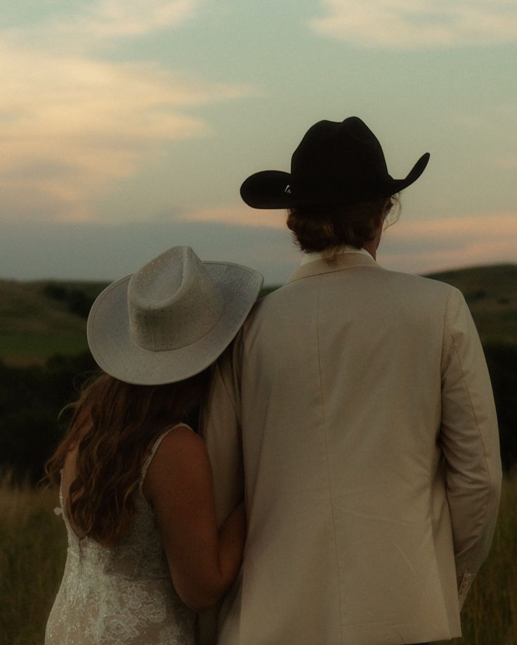 a man and woman in cowboy hats looking out at the sunset on their wedding day