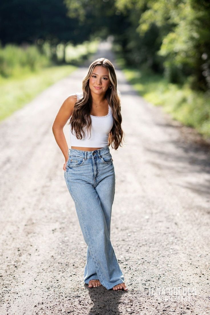 a woman standing in the middle of a dirt road wearing high waist jeans and a white crop top