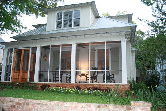 a white house with screened porches and chairs on the front lawn, next to a brick wall