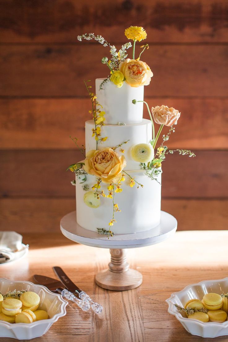 a three tiered white cake with yellow flowers on top and other desserts around it