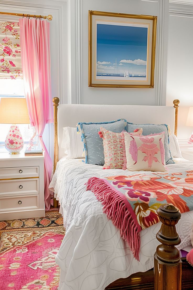 a white bed with pink and blue pillows in a bedroom next to a window filled with curtains