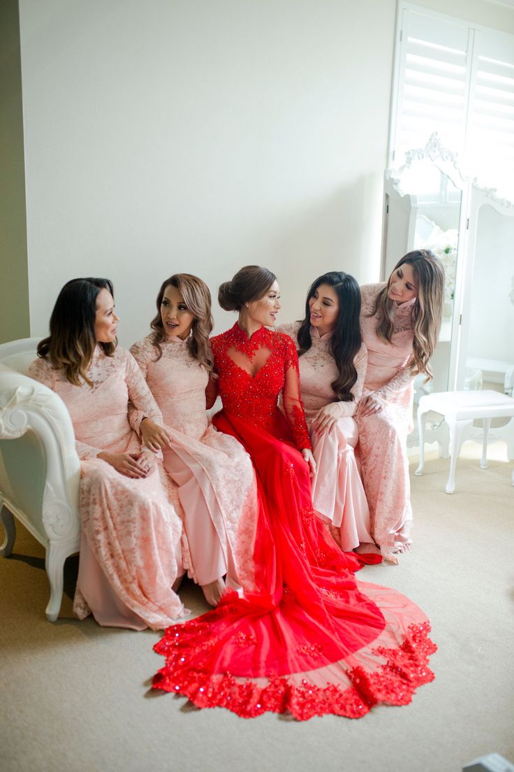 four women in pink dresses are sitting on a white couch and one is wearing a red dress