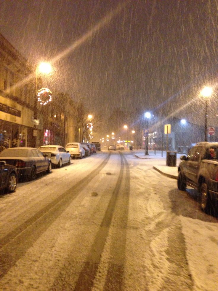 cars parked on the side of a snowy street at night with lights in the background