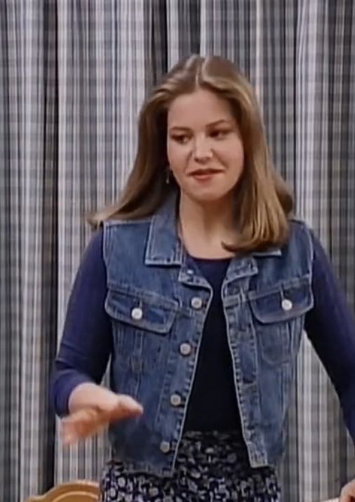 a woman standing in front of a window with her hands out to the side while wearing a jean jacket