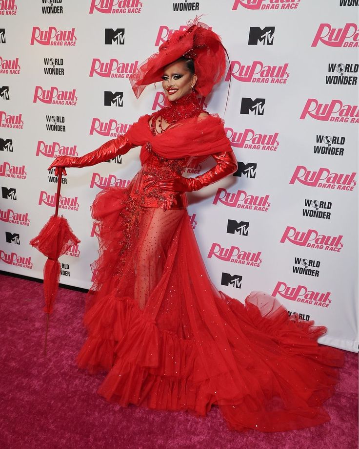 a woman in a red dress and hat posing on the pink carpet at an event