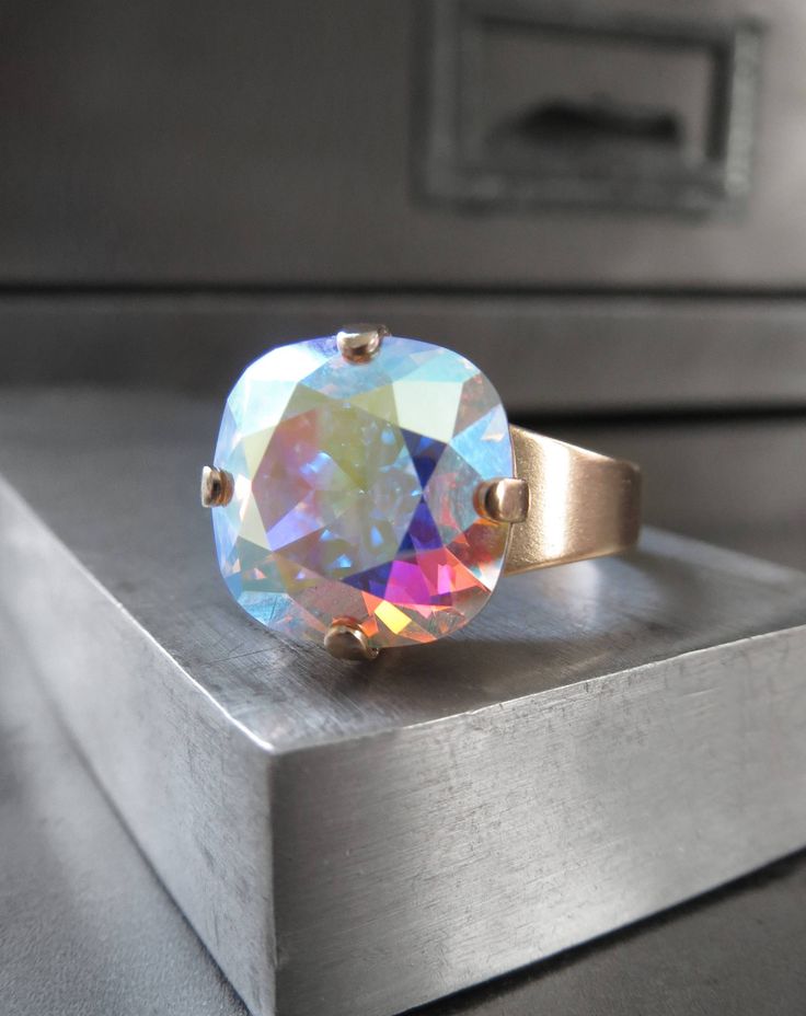 Full of iridescent shimmer & sparkle! Renowned for impeccable faceting and quality, this 'Crystal AB' Swarovski crystal flashes playfully with soft, pastel hues in all hues of the rainbow! The square cushion-cut crystal is set into a matte, soft gold-plated bezel on an adjustable ring band. Fits sizes approx. 6 to 9 (may not fit smaller fingers). Also available with Antiqued Silver adjustable ring band - sold separately. Cushion-cut Swarovski crystal: 1/2" x 1/2" (12 x 12mm) Comfortable and