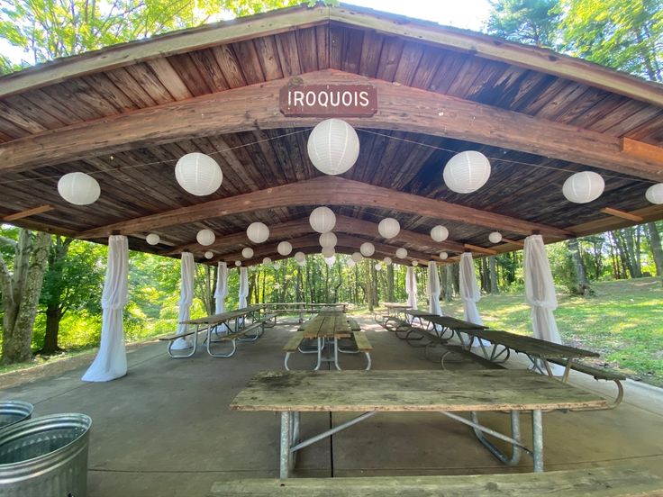 an outdoor covered picnic area with tables and lanterns hanging from the roof, surrounded by trees