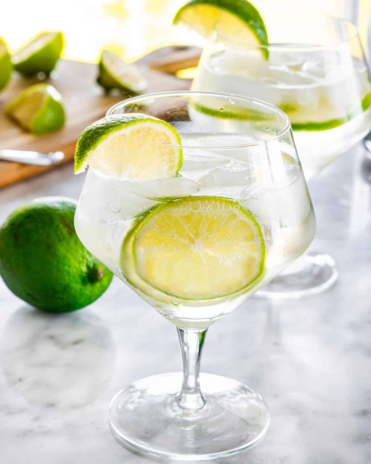 two glasses filled with lemonade and limes on a counter top next to sliced limes