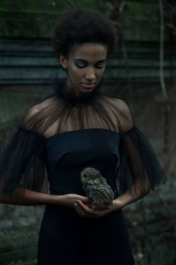 a woman with an owl in her hand is wearing a black dress and standing outside