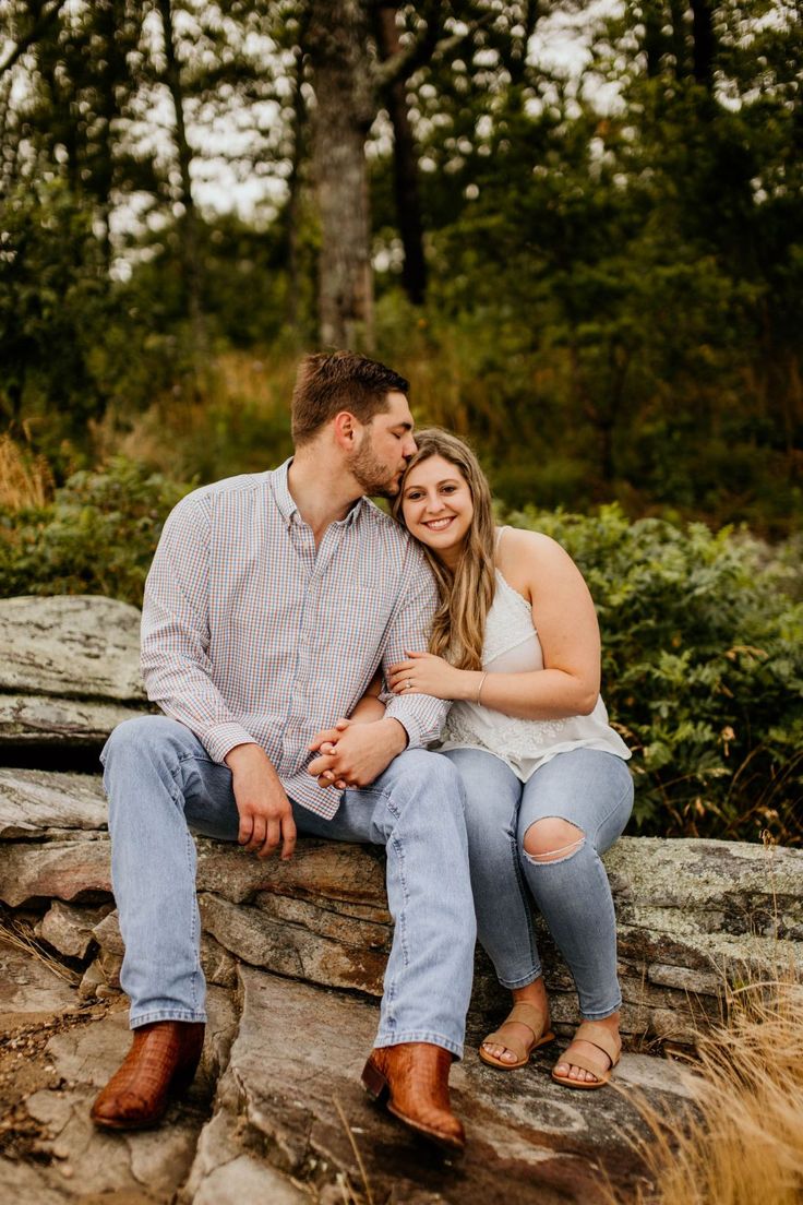 a man and woman sitting on top of a rock in front of some trees with their arms around each other