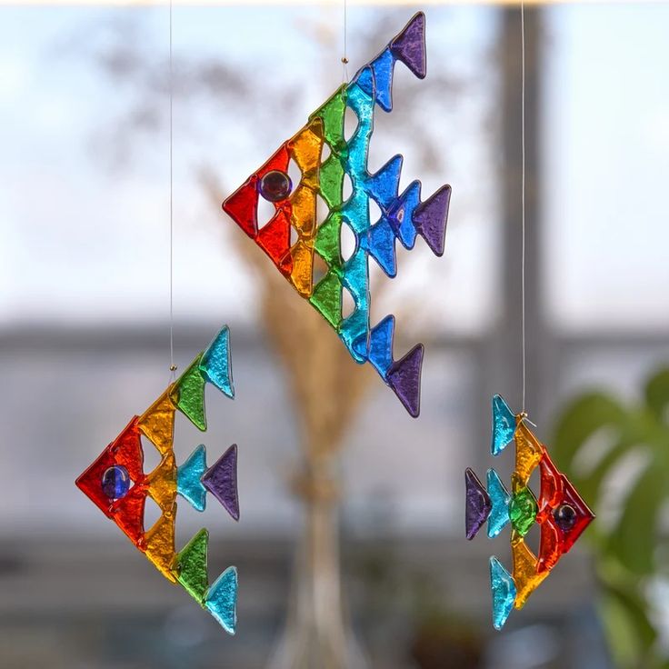 three colorful glass fish hanging from strings in front of a vase filled with flowers and greenery