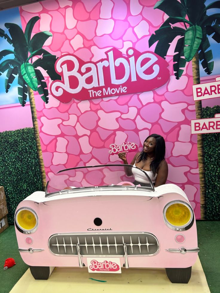 a woman sitting on top of a pink car in front of a barbie movie sign