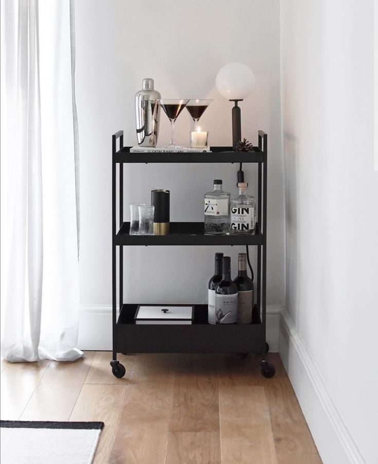 a black shelf with bottles and glasses on it in front of a white wall next to a window