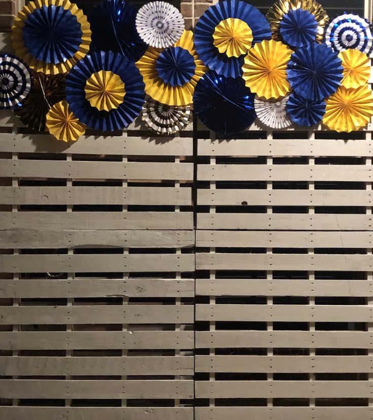 several blue and yellow paper fans are hanging on the side of a wall with wood pallets