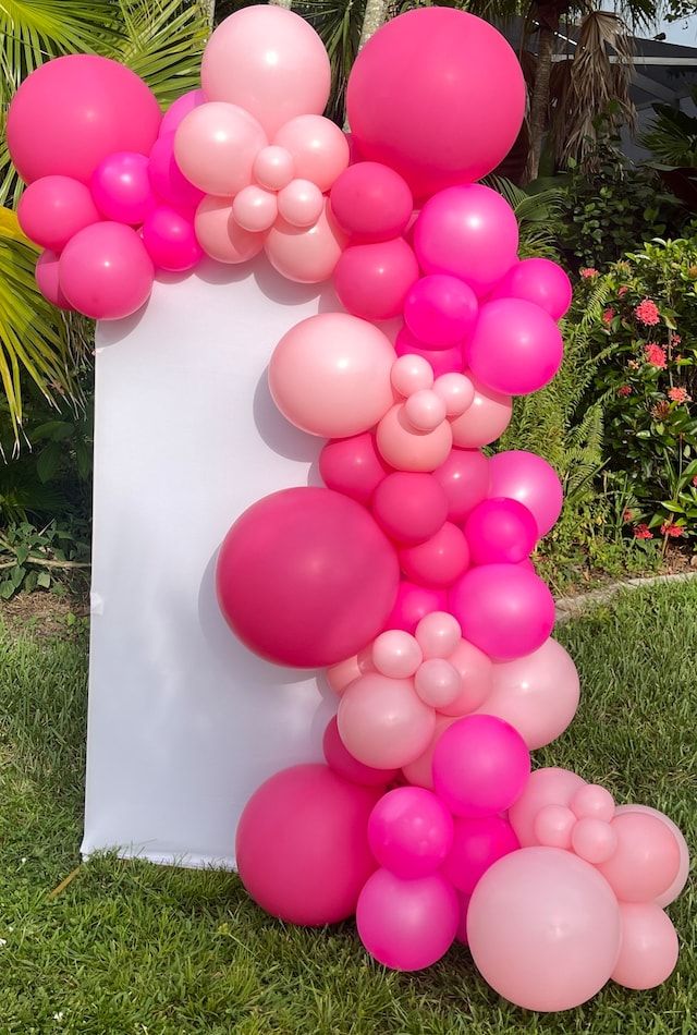 a bunch of pink balloons are hanging from a white sign in the grass with palm trees behind it