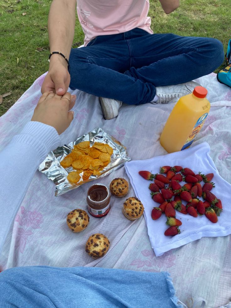 two people sitting on a blanket with food and drinks in front of them, one holding the other's hand