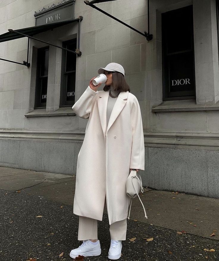 a woman standing on the street drinking out of a cup while wearing a white hat and coat