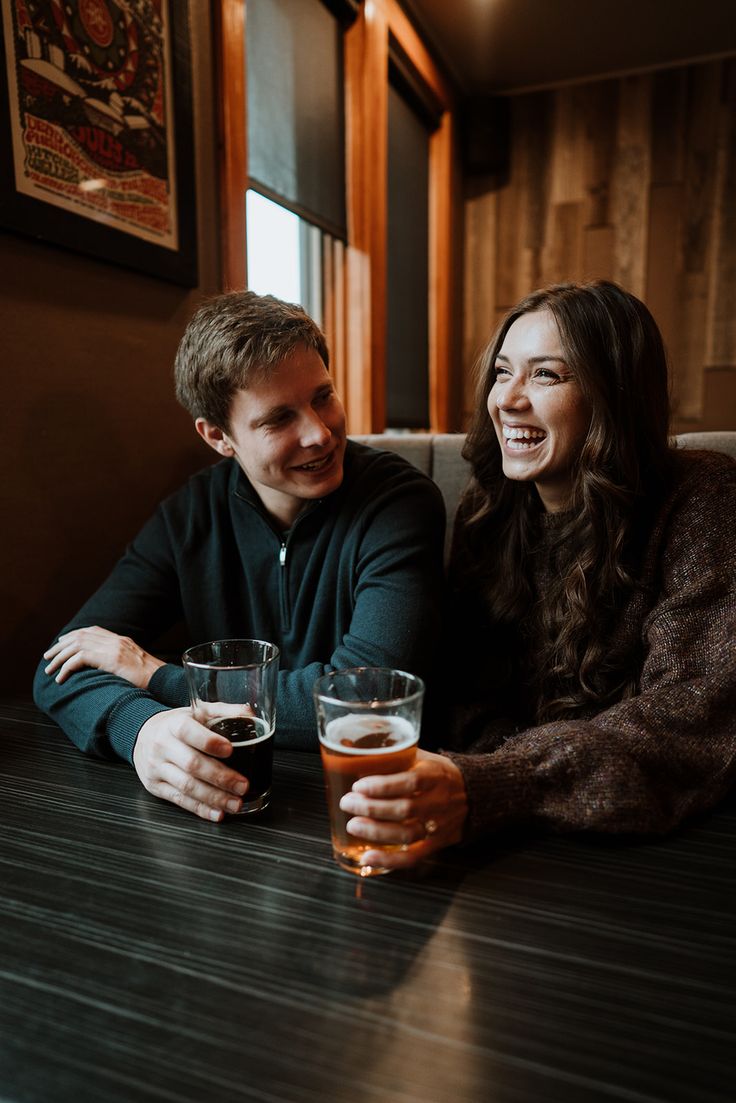 a man and woman sitting at a table with two beers in their hands, smiling