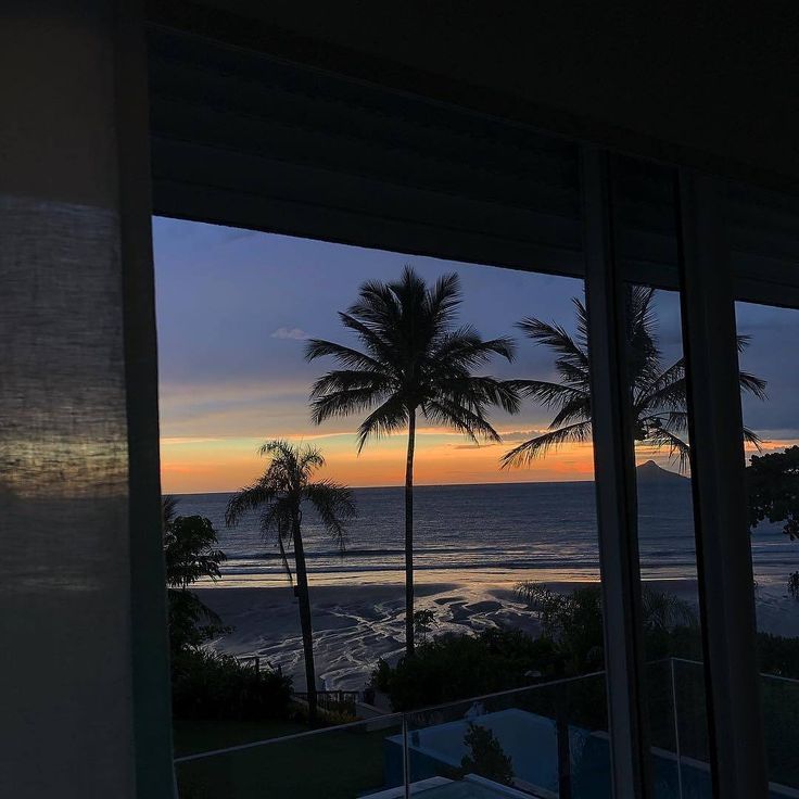 the sun is setting over the ocean and palm trees are seen through large glass windows