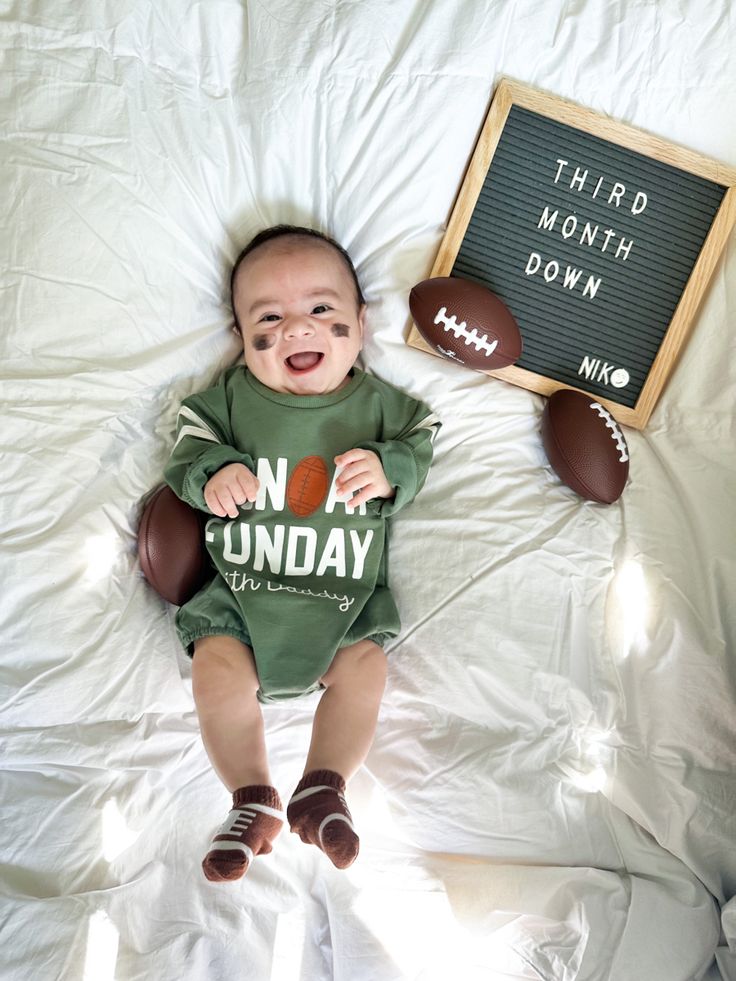 a baby laying on a bed next to a sign that says third month down with footballs