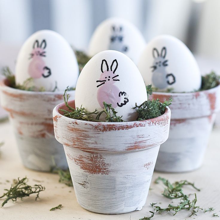 four decorated eggs with faces drawn on them sitting in small white pots filled with grass