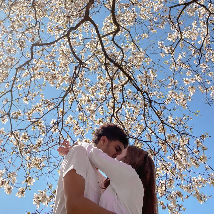 a man and woman kissing under a tree with white flowers in the sky behind them