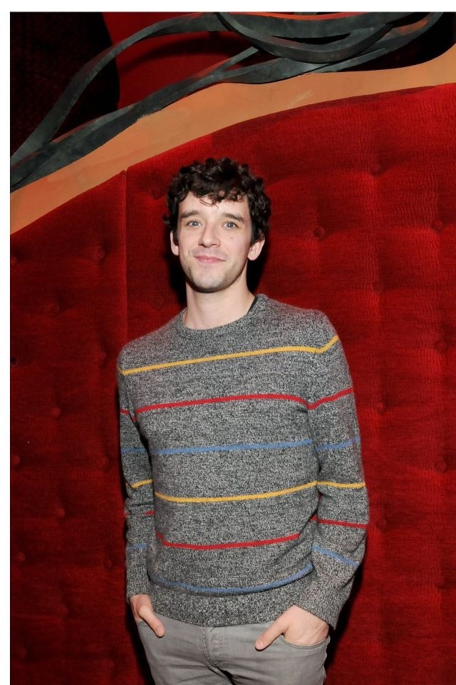 a man standing in front of a red wall wearing a striped sweater and grey pants