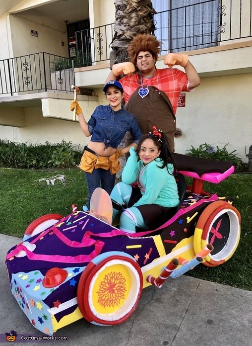 three children are sitting on a toy car in front of a building with balconies