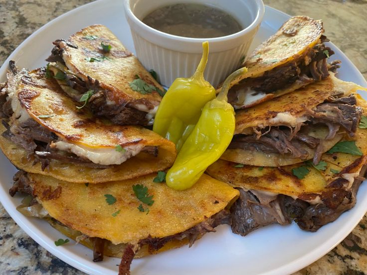 a white plate topped with tortillas covered in meat and cheese next to a cup of sauce