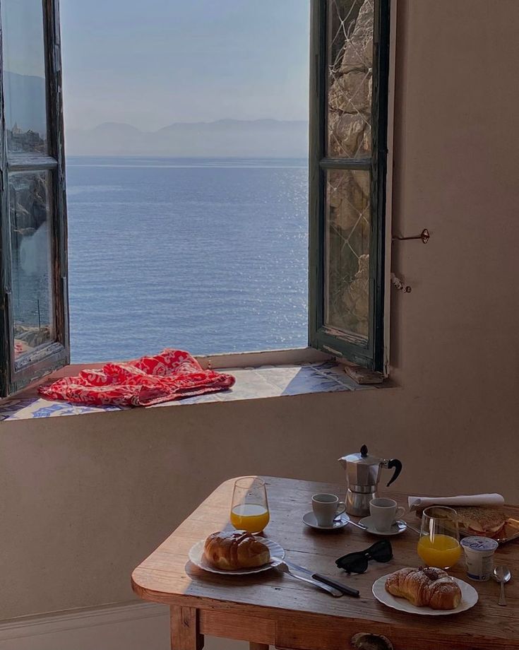 a table with breakfast food on it in front of an open window overlooking the ocean