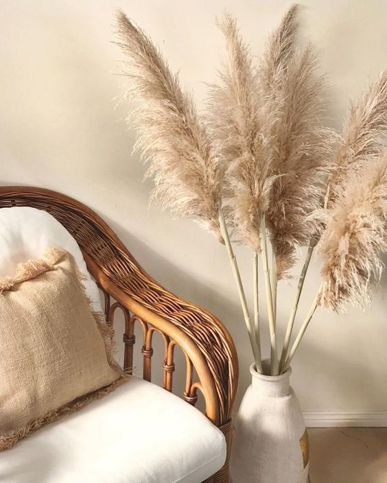 a white vase filled with dry grass next to a wicker chair and throw pillow