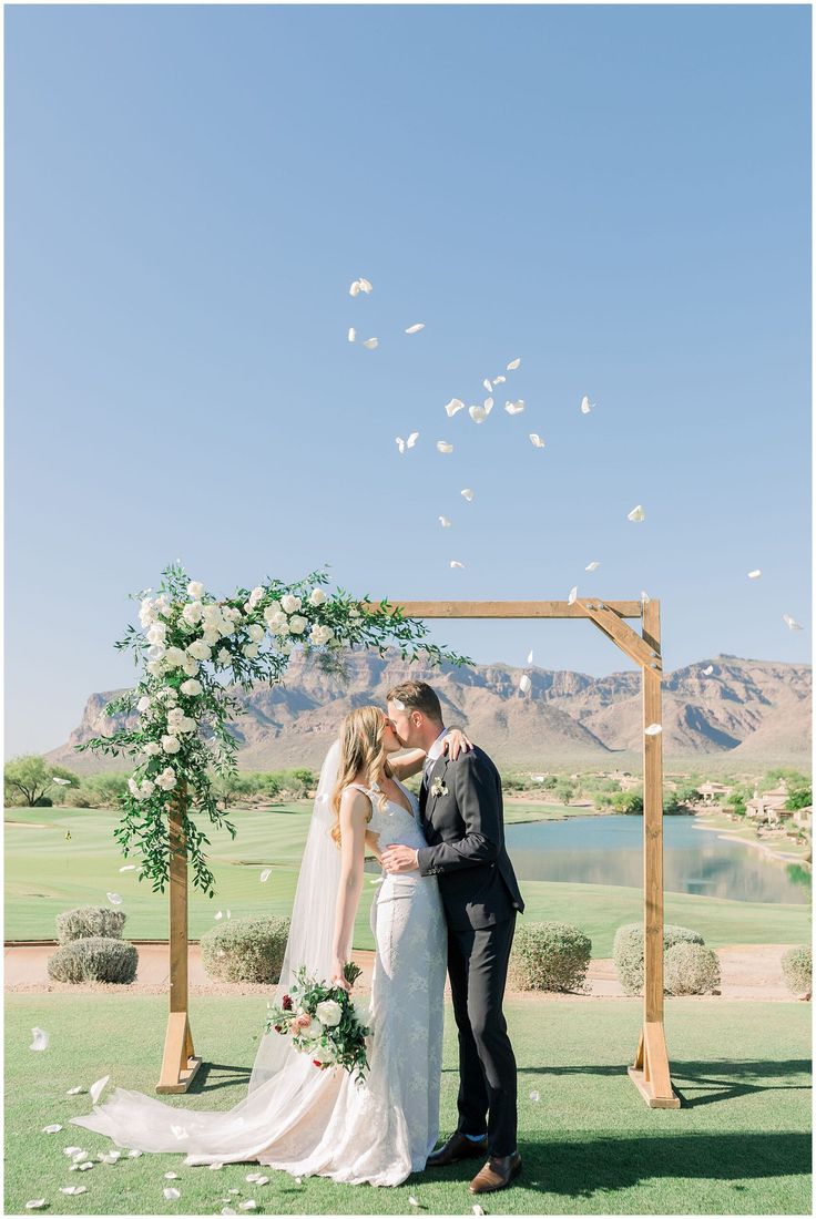 a bride and groom kissing under an arch with white flowers in front of the mountains