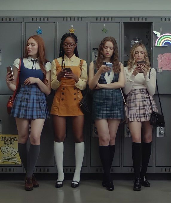 four girls standing in front of lockers looking at their cell phones while wearing school uniforms