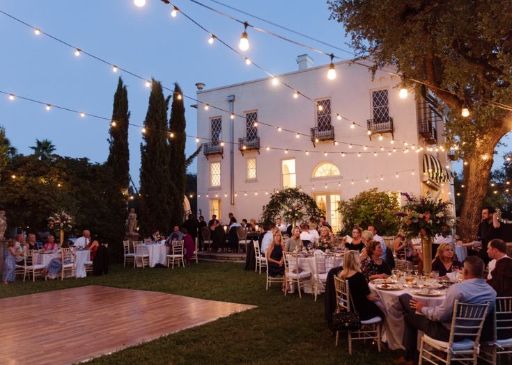 people sitting at tables in front of a building with lights strung from the roof and trees