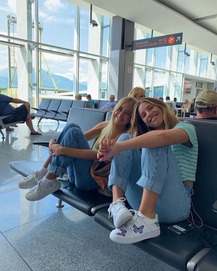 two young women sitting on the back of an airport luggage cart smiling at the camera