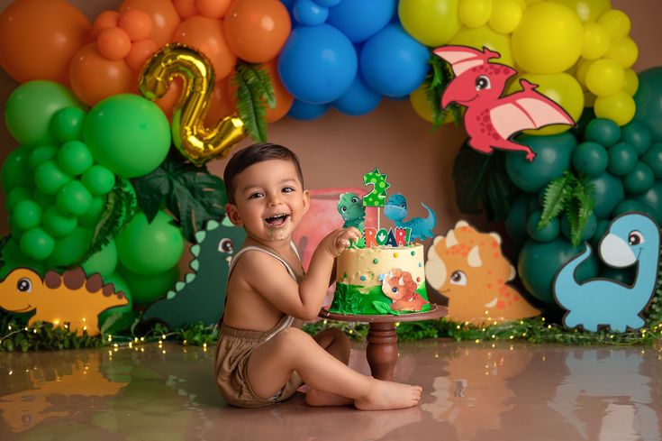 a little boy sitting on the floor with a cake in front of him and balloons behind him